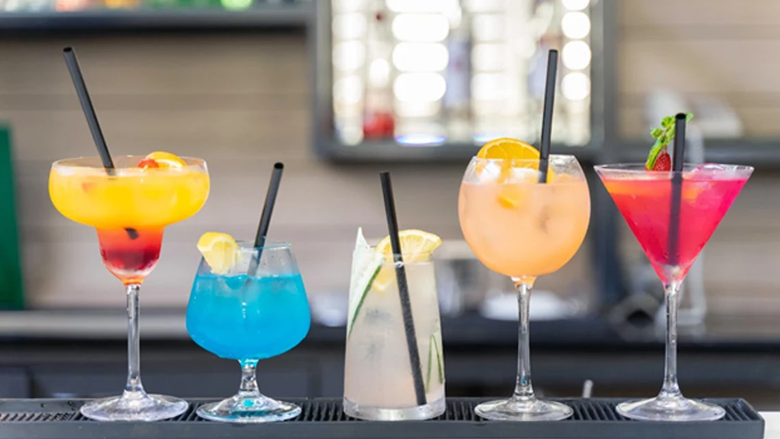 An array of cocktail glasses with fruit slices, black straws, and adaptogen drinks of various colors
