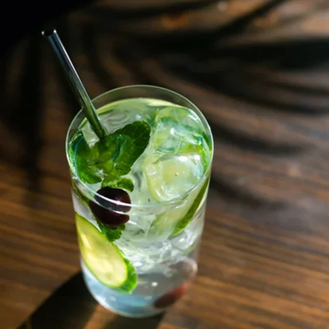 A glass of cannabis-infused spritzer with a straw, ice cubes, mint leaves, and cucumber slices